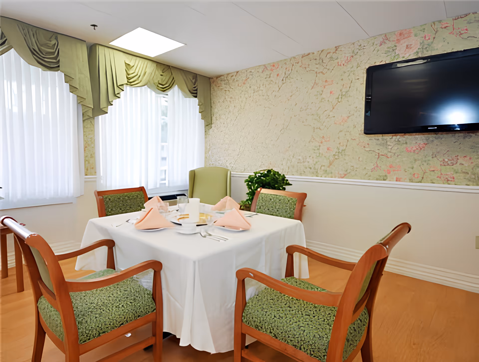 A dining area with a square table covered with a white tablecloth, set with pink folded napkins, plates, and utensils. Four wooden chairs with green patterned cushions surround the table. The room has large windows with sheer white curtains and green valances. A flat-screen TV is mounted on a wall with a floral textured wallpaper, and a green armchair and a potted plant are visible in the corner.