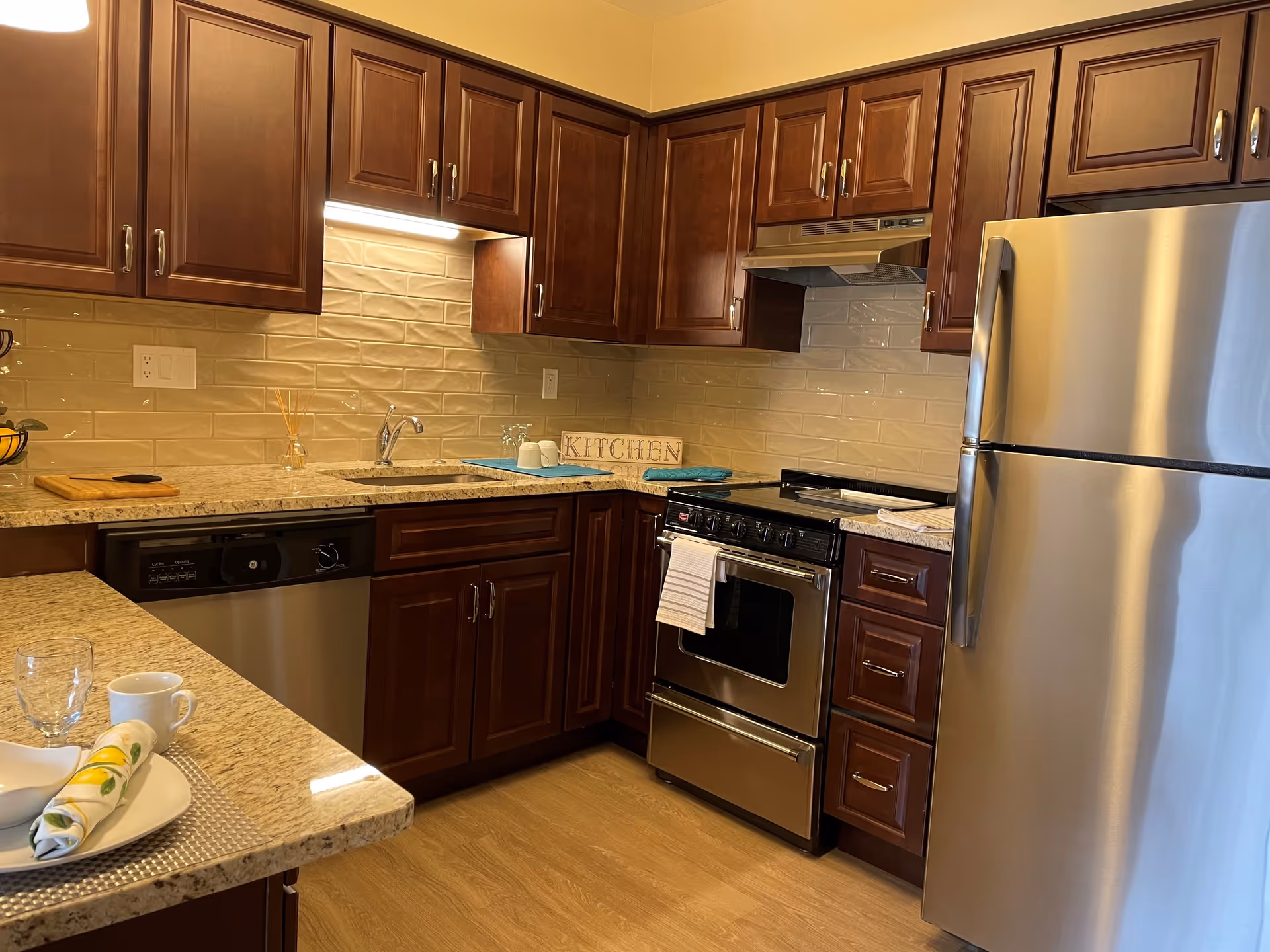 A modern kitchen with dark wooden cabinets, beige tiled backsplash, granite countertops, stainless steel refrigerator, stove, and dishwasher. There is a small sign on the counter that reads 'KITCHEN' and a place setting with a plate, napkin, cup, and glass on the countertop.