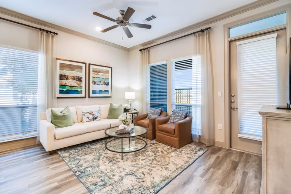 A bright living room with a white sofa adorned with green and patterned pillows, two brown leather armchairs, a round glass coffee table on a patterned area rug, two abstract paintings on the wall, large windows with sheer curtains, a ceiling fan, and a door with blinds.