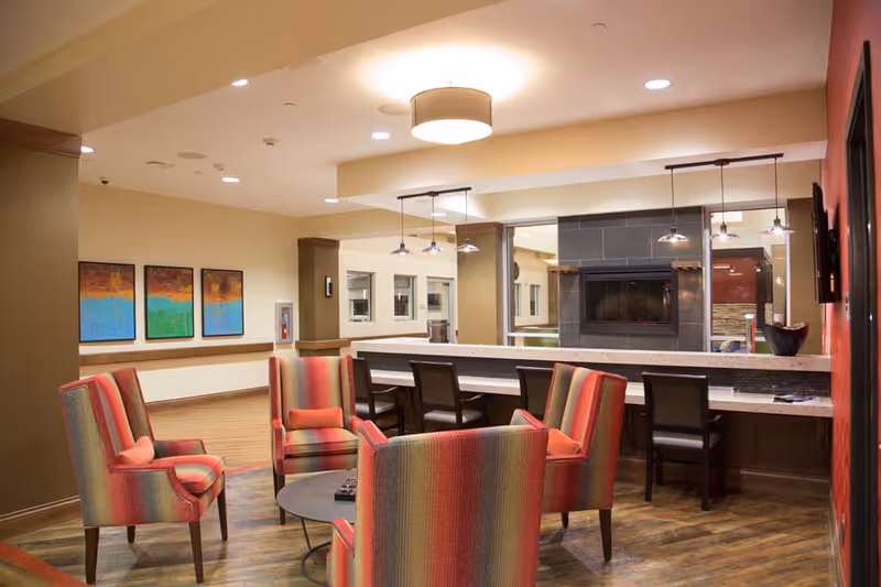 A cozy common area with four striped armchairs arranged around a small round table. Behind the chairs is a long counter with several chairs, and a modern fireplace set in a dark tiled wall. The room has warm lighting, wooden flooring, and colorful abstract paintings on the wall.