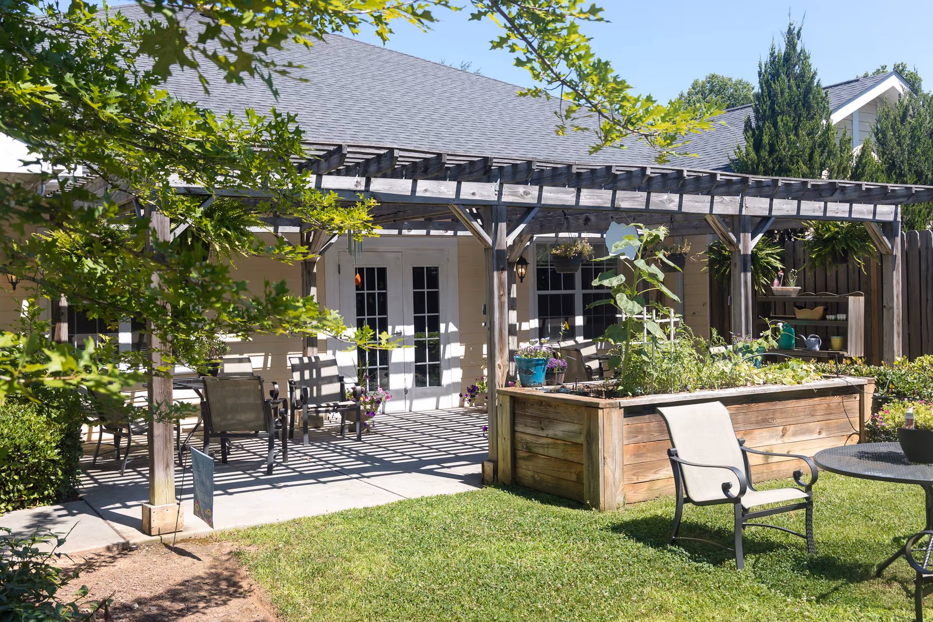 Shaded outdoor patio with a wooden pergola, chairs, a raised garden bed and lawn in front of a building.