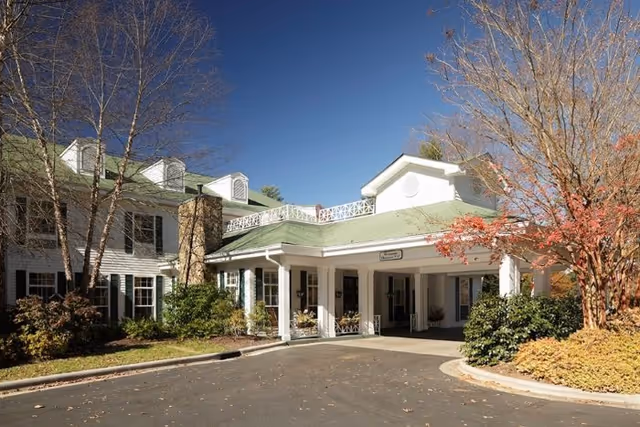 Front entrance of a white senior living building with a covered porte-cochère, green roof, and landscaped trees under a clear blue sky.