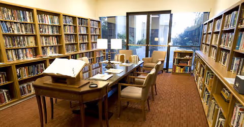 A small library or reading room with bookshelves along both walls, a long wooden table with chairs and lamps, and glass doors/windows at the far end.