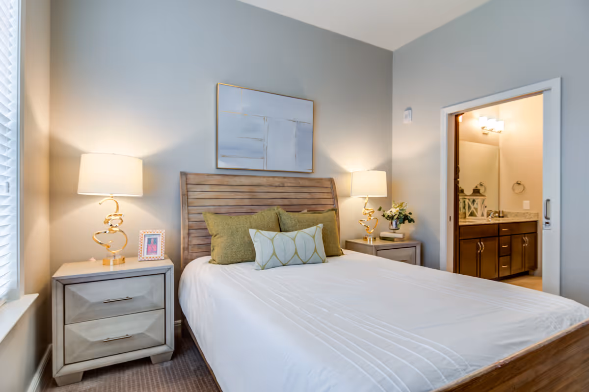 A bedroom with a wooden bed frame and white bedding, accented by three green pillows. There are two matching nightstands on either side of the bed, each with a modern gold lamp. A framed abstract painting hangs above the bed. To the right, an open doorway reveals a bathroom with a vanity, mirror, and lighting fixtures.