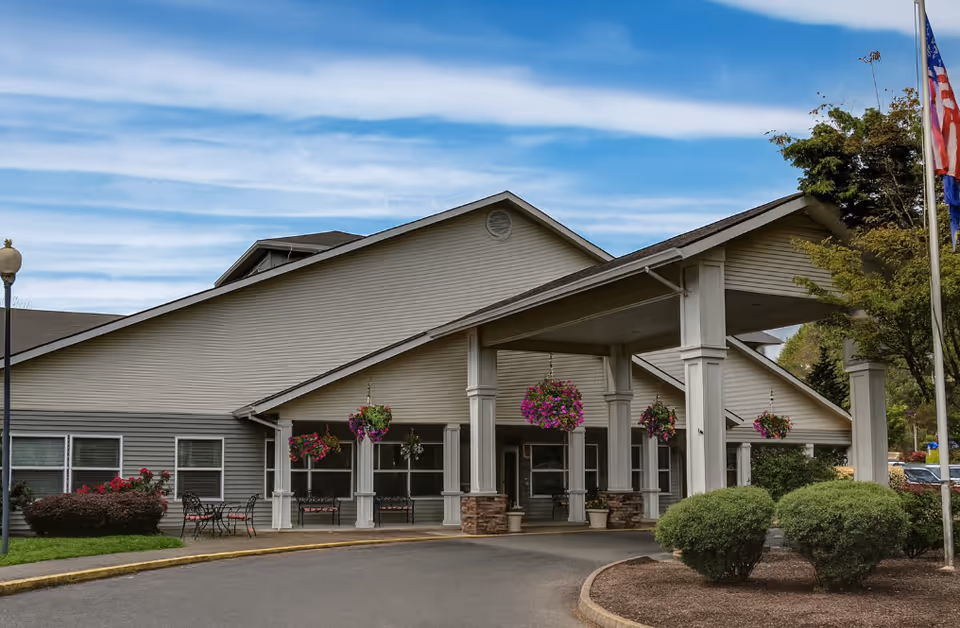 Covered front entrance of a senior living building with columns, hanging flower baskets, and a curved driveway.