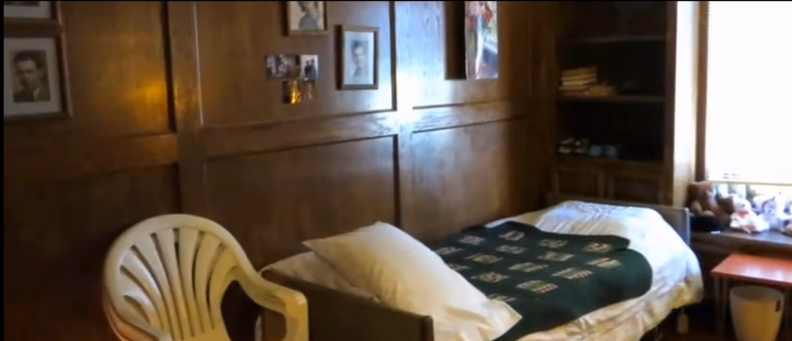 A small bedroom with wood-paneled walls featuring several framed pictures. The room contains a single bed with white sheets and a dark green patterned blanket, a white plastic chair, a wooden shelf with folded clothes and shoes, and a window with stuffed animals on the windowsill. A small wooden table and a waste bin are also visible near the window.