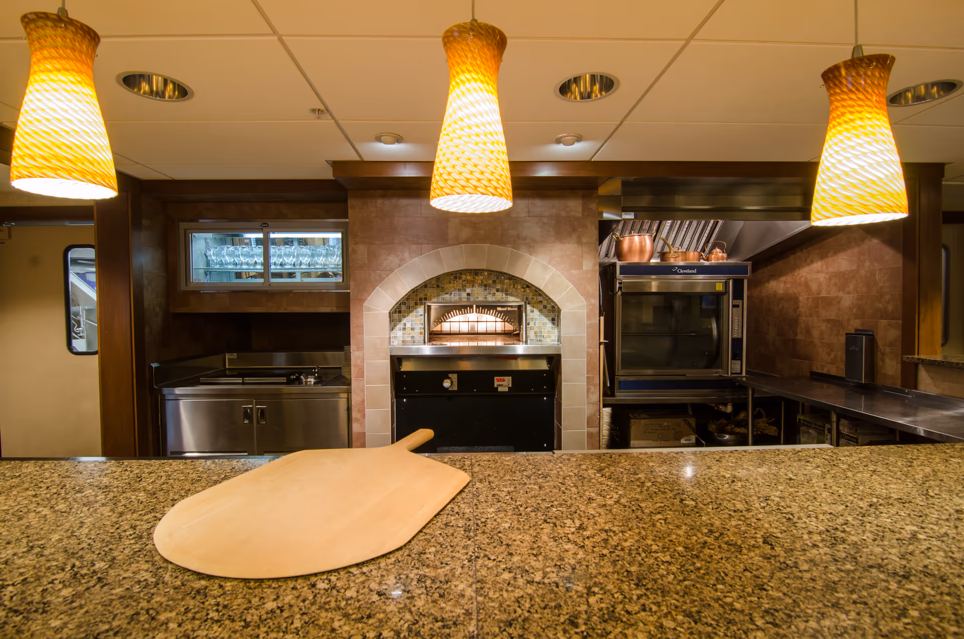 Interior view of a kitchen area with a granite countertop in the foreground, a wooden pizza peel resting on the counter, three hanging yellow pendant lights above, a built-in oven with a tiled arch surround in the center, and stainless steel appliances and shelves in the background.