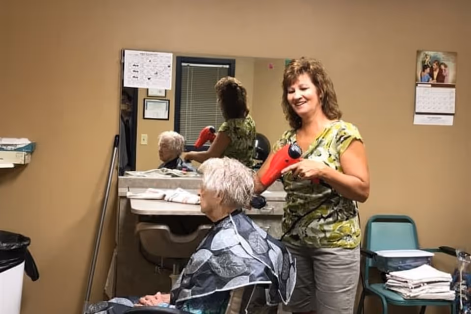 A woman is blow-drying the hair of an elderly person seated in a chair in a small room with beige walls. There is a large mirror on the wall reflecting both individuals. The room contains a calendar, a mop, a trash can, and a chair with folded towels on it.