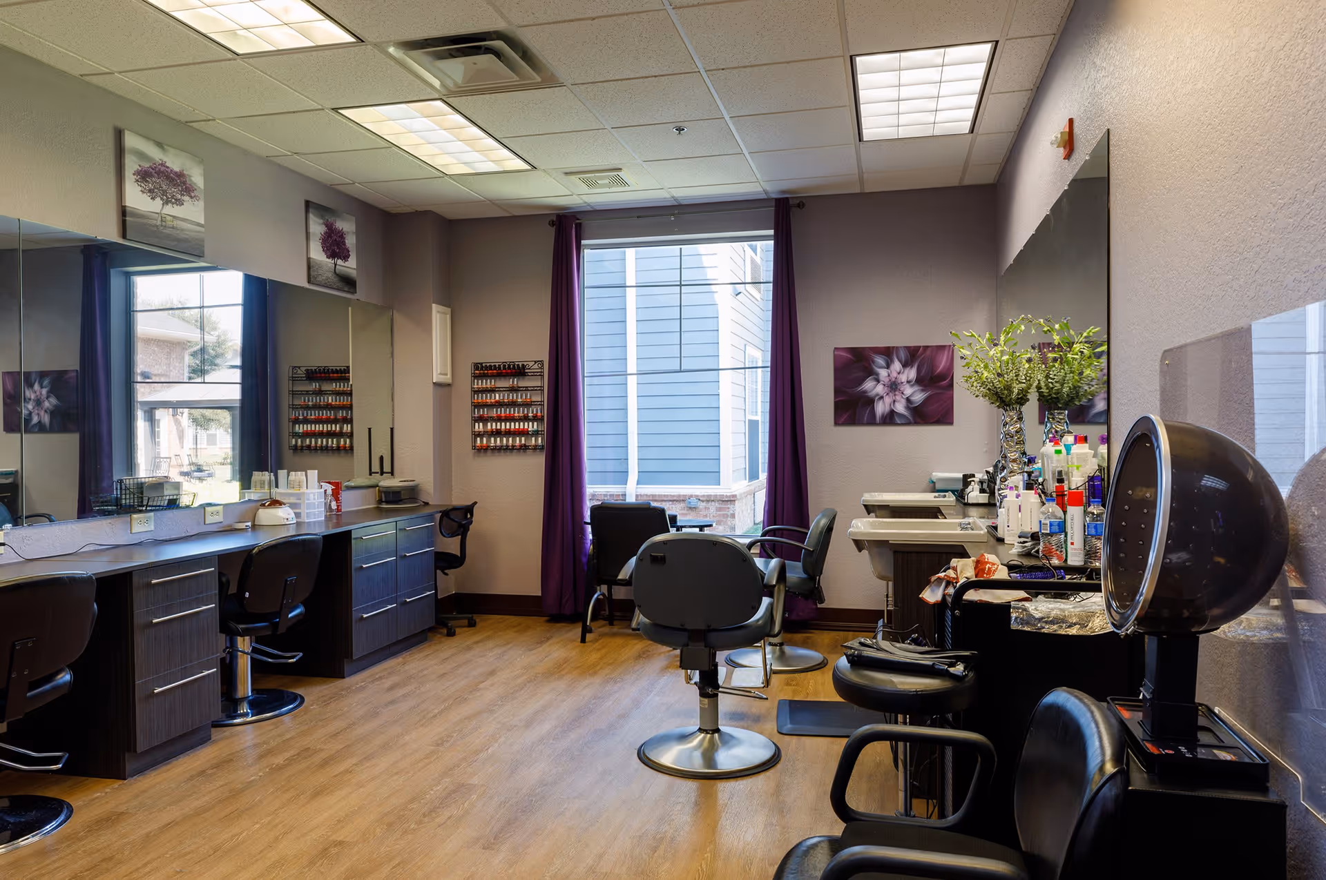 Interior view of a salon area in a senior living facility with multiple styling chairs, large mirrors on the walls, nail polish racks, a hair dryer chair, and a window with purple curtains letting in natural light.