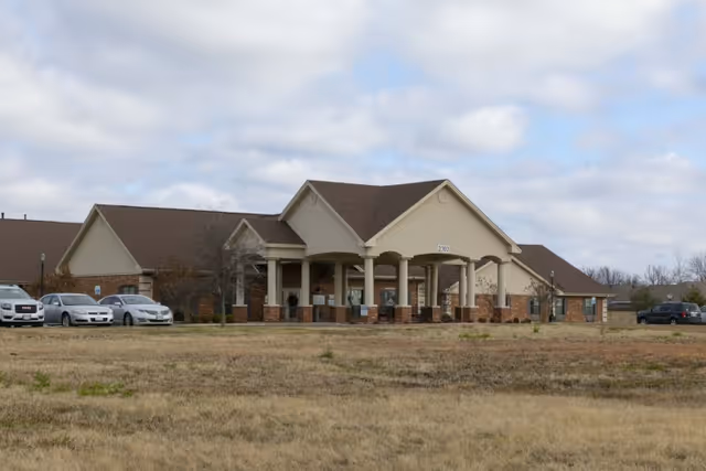 Front exterior of a single-story senior living facility with a covered columned entrance, parked cars, and a grassy foreground.