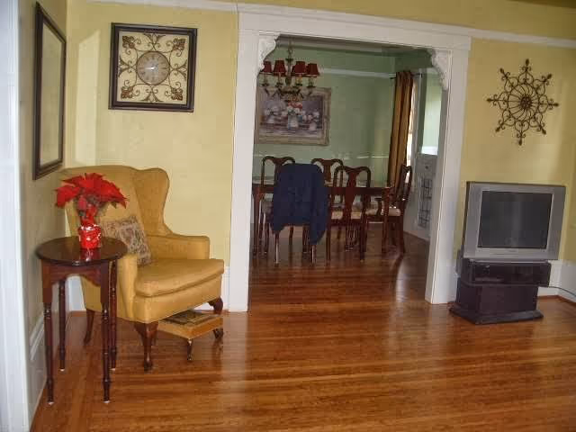 Interior view of a senior living facility showing a cozy living room with a yellow armchair next to a small wooden side table with a red poinsettia plant. The room has wooden flooring and light yellow walls. Through an open doorway with white trim, a dining room with a wooden dining table and chairs is visible, with a chandelier hanging above the table and a framed painting on the green wall.