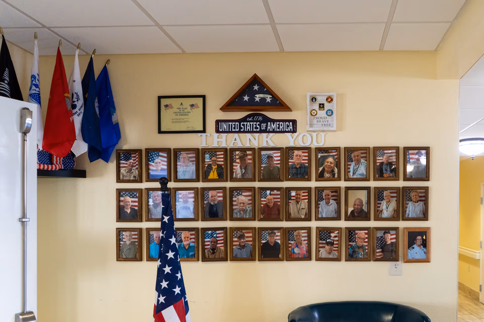 Wall display featuring framed photos of veterans with American flags in the background, a folded American flag in a triangular case, various military flags, and plaques with patriotic messages including 'Thank You' and 'United States of America'.