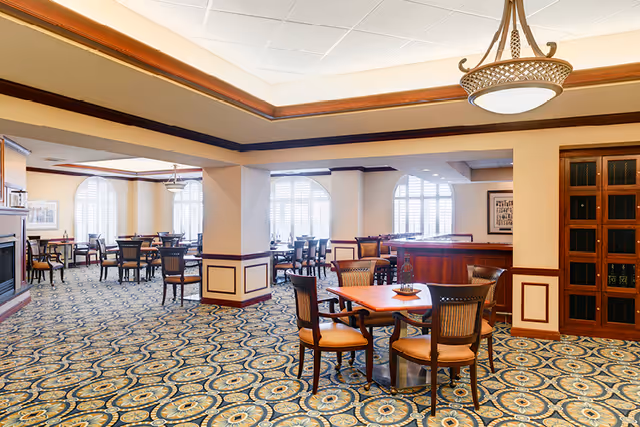 Spacious dining room with several tables and chairs, patterned carpet, large arched windows, and warm wood trim.