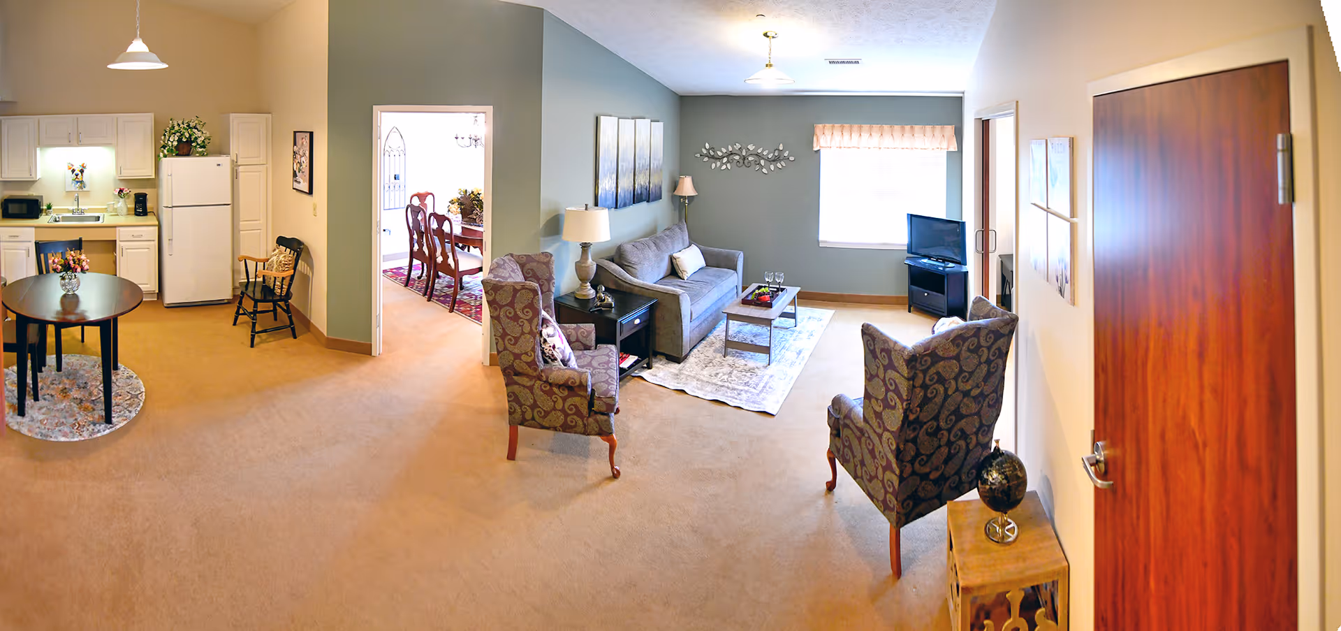 A spacious and well-lit living area in a senior living facility featuring a small kitchen with white cabinets, a refrigerator, and a round dining table with chairs. Adjacent to the kitchen is a cozy living room with a sofa, two patterned armchairs, a coffee table, a TV on a stand, and decorative wall art. A doorway leads to a formal dining room with a wooden dining table and chairs. The room has beige carpeting and soft lighting from ceiling fixtures and lamps.