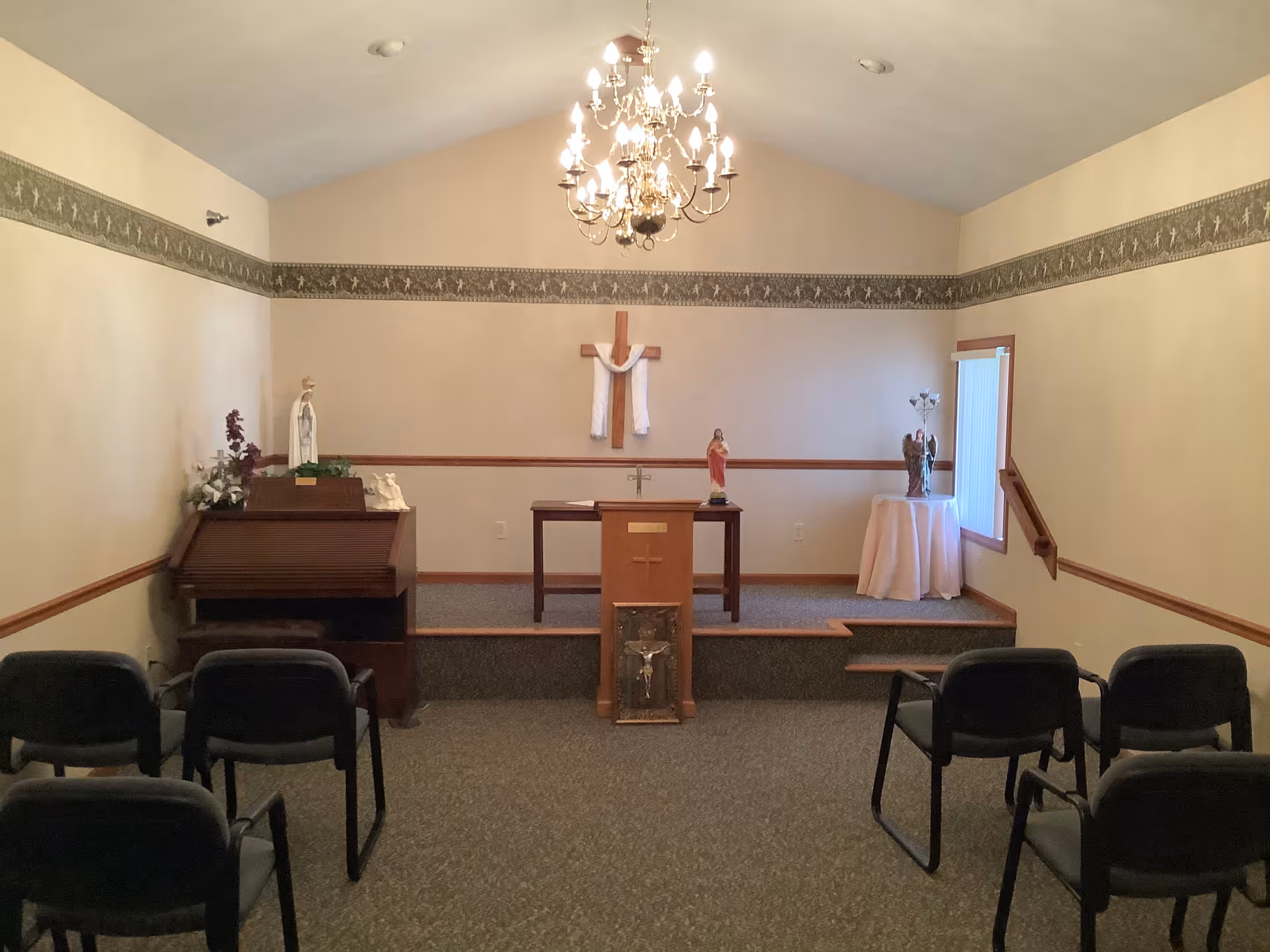 Small chapel-like room with an altar and wooden cross, religious statues, rows of chairs, and a chandelier.