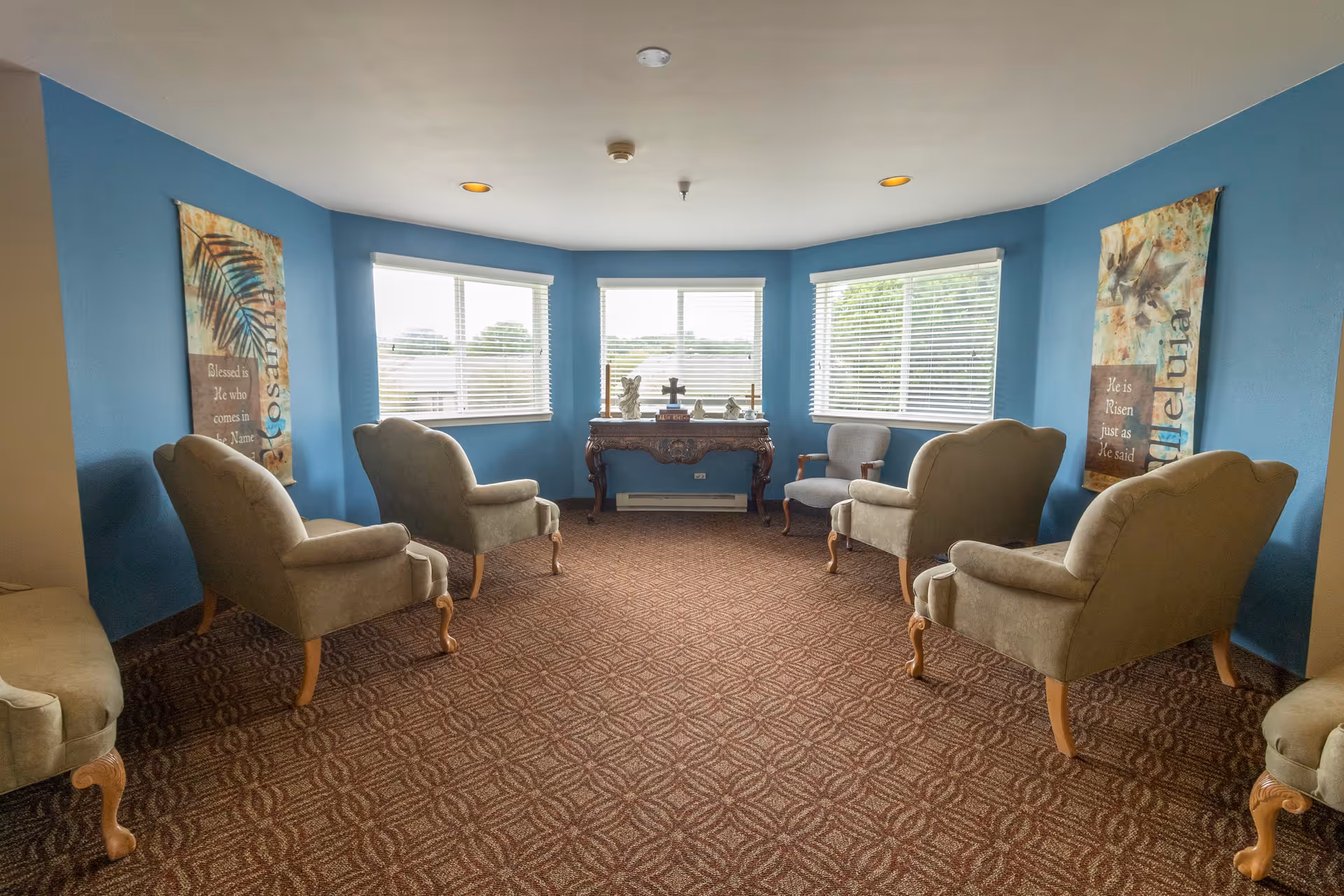 A small sitting room with blue walls and three large windows letting in natural light. The room contains six upholstered armchairs arranged in a semi-circle facing a wooden table with decorative items including crosses. Two vertical banners with religious text hang on the walls.