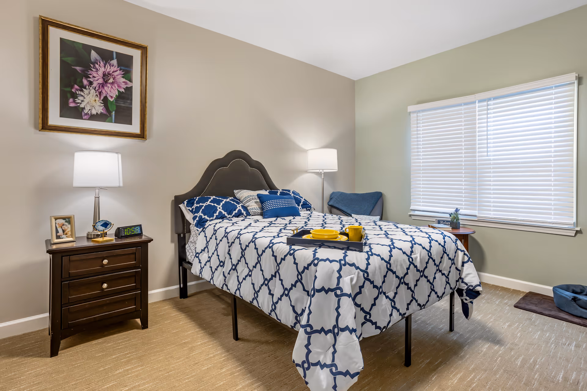A neatly made bed with a white and blue patterned bedspread in a bedroom. There are several pillows on the bed and a tray with yellow dishes and a mug placed on top. To the left of the bed is a dark wooden nightstand with a lamp, a framed photo, a decorative item, and a digital clock showing 10:58. On the wall above the nightstand is a framed picture of flowers. To the right of the bed is a window with white blinds, a small round table with a plant and a 'Relax' sign, and a blue chair with a blanket draped over it. The floor is carpeted and there is a pet bed in the corner.