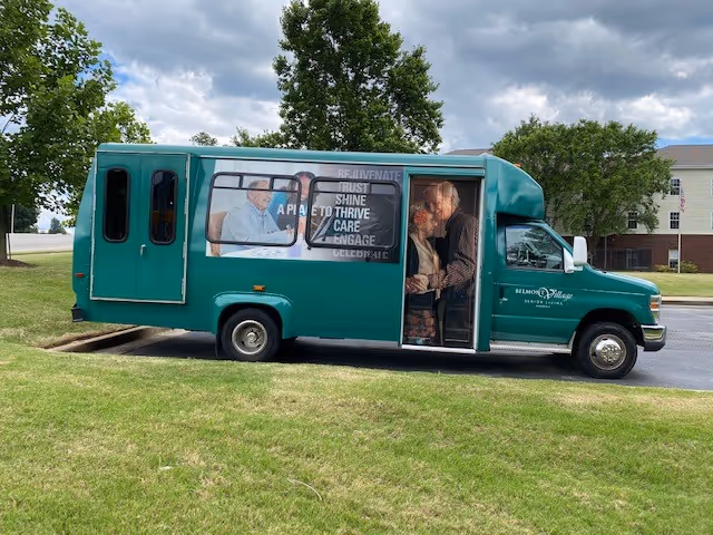 Teal Belmont Village shuttle bus with open door parked on grass near buildings.
