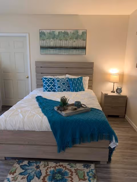 A bedroom with a wooden bed frame and headboard, a white comforter, and three decorative blue and white pillows. A teal throw blanket is draped across the foot of the bed with a tray holding decorative items. To the right of the bed is a wooden nightstand with a lamp and a small clock. A door is visible on the left wall, and a painting of trees hangs above the headboard. The floor has a floral patterned rug near the bed.