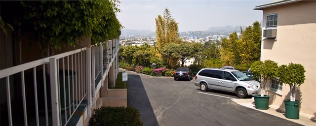 Parking area outside a residential building with two parked cars, potted plants, and a view of trees and distant hills under a clear sky.