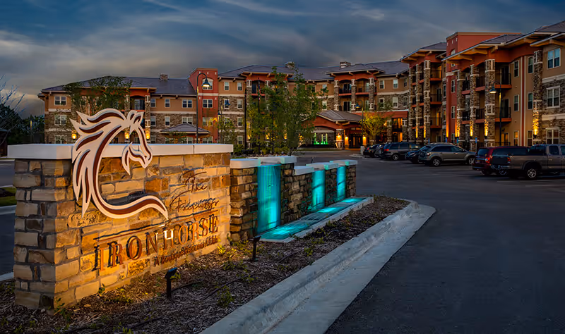 Illuminated stone entrance sign and water features in front of the Ironhorse senior living building at dusk.