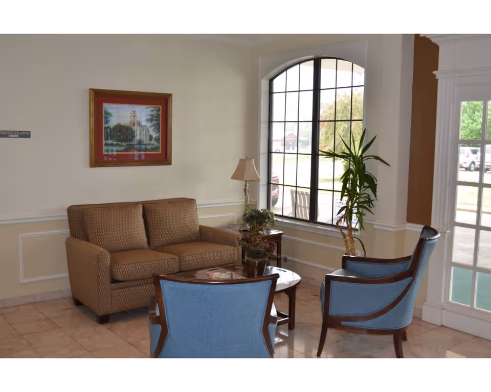 A cozy seating area inside a facility with a brown loveseat, two blue armchairs, a wooden coffee table with plants, a floor lamp, a large window with black grid frames, and a framed painting on the wall.