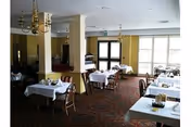 Interior view of a dining room with multiple tables covered with white tablecloths, wooden chairs, chandeliers hanging from the ceiling, and large windows allowing natural light to enter.