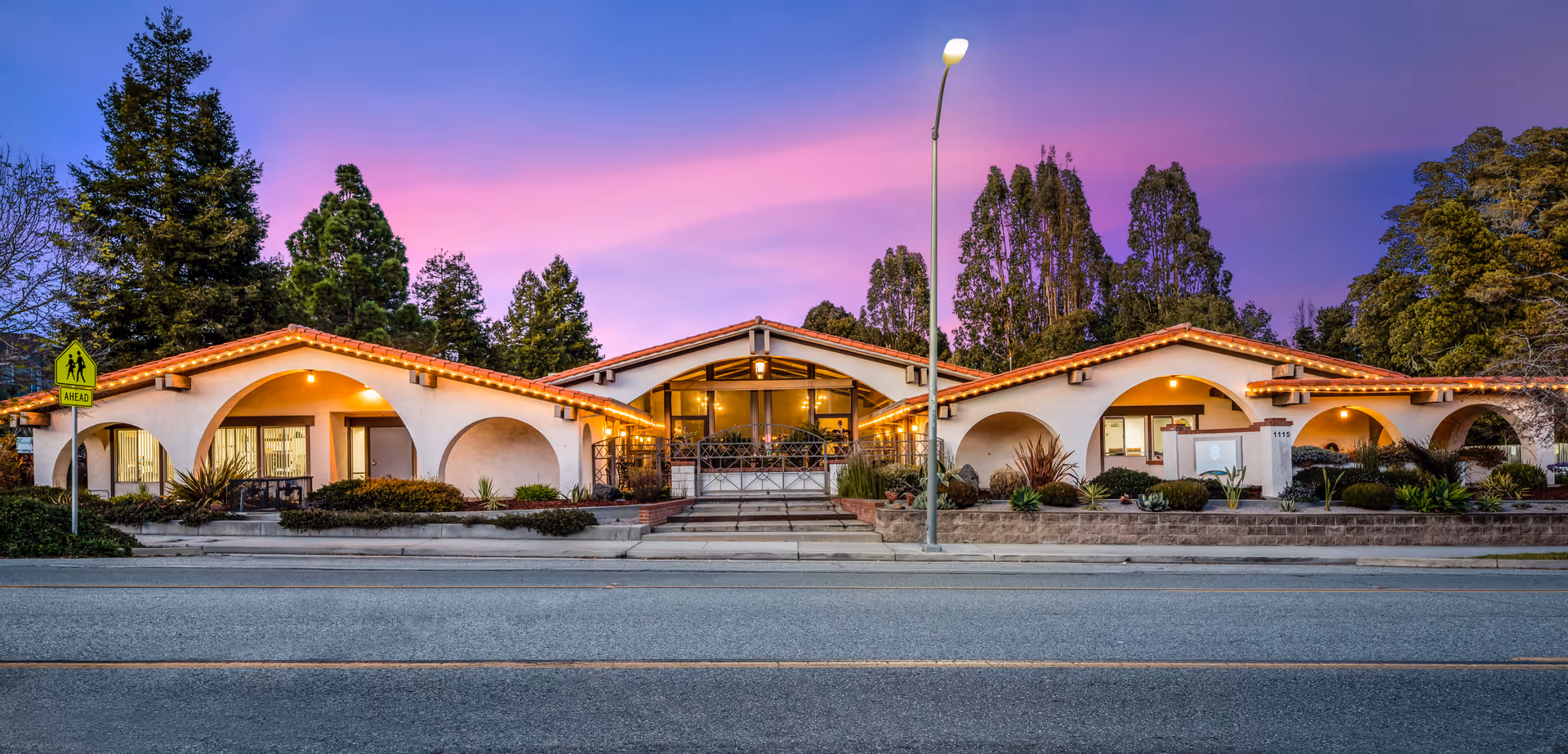 Exterior view of Santa Cruz Post Acute facility at dusk with a colorful purple and pink sky. The building features a Spanish-style architecture with white walls, red tile roofs, and arched entryways. There are well-maintained shrubs and plants in front, a street with a pedestrian crossing sign, and a streetlamp in the foreground.
