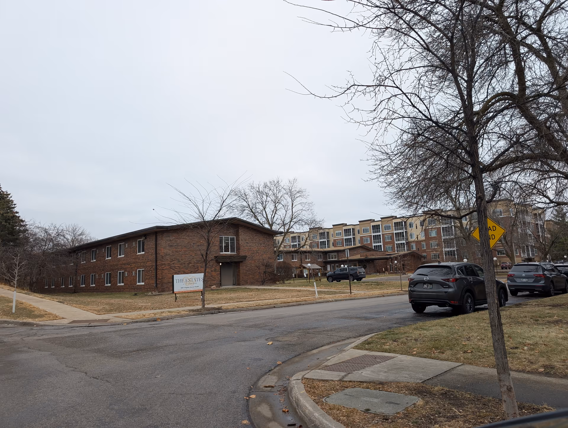 Exterior view of The Estates at St. Louis Park facility showing a brick building on the left with a sign in front, a larger multi-story building in the background, parked cars along the street, leafless trees, and a cloudy sky.