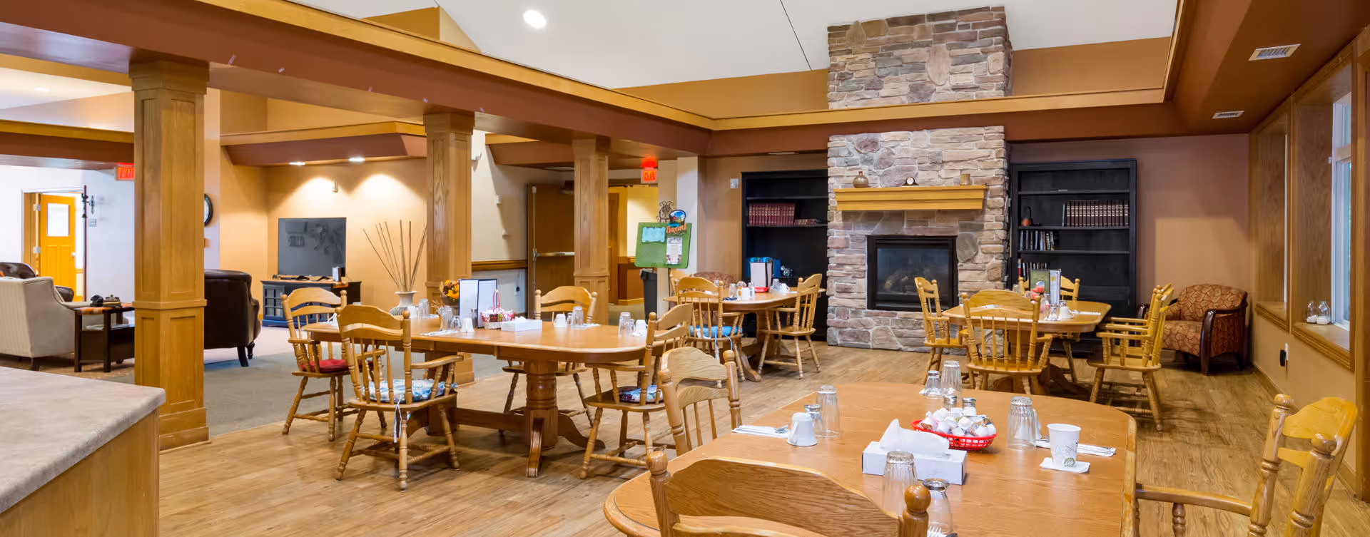 A cozy dining area in a senior living facility featuring wooden tables and chairs arranged around the room. The space includes a stone fireplace with a wooden mantel, bookshelves, and warm lighting. The room has wooden beams and columns, with a mix of carpet and wood flooring, creating a welcoming atmosphere.