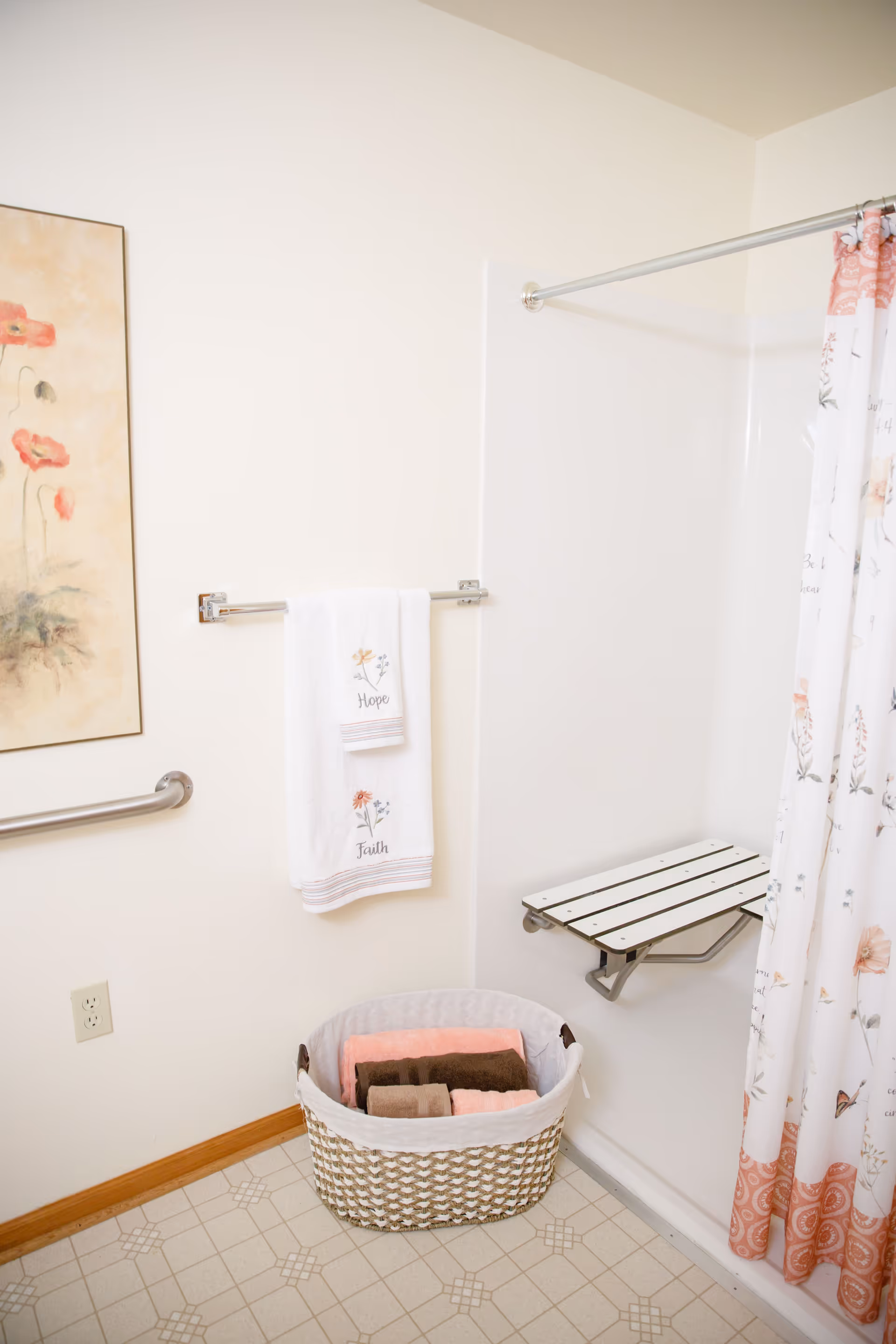 A bathroom corner featuring a white wall with a floral painting, a towel rack holding two white towels embroidered with the words 'Hope' and 'Faith', a woven basket on the floor containing folded towels in pink and brown, a foldable shower seat attached to the shower wall, and a floral shower curtain with pink and orange accents.