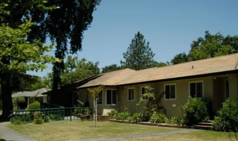 Single-story beige care home with front yard, trees, and walkway.