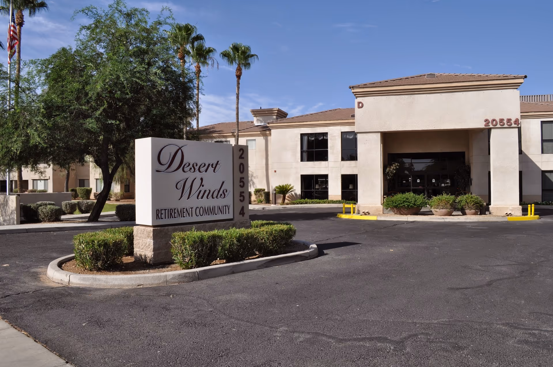 Exterior view of Desert Winds Retirement Community building with a large sign displaying the community name and address 20554. The building is two stories with beige walls and a covered entrance. There are palm trees and shrubs around the property under a clear blue sky.