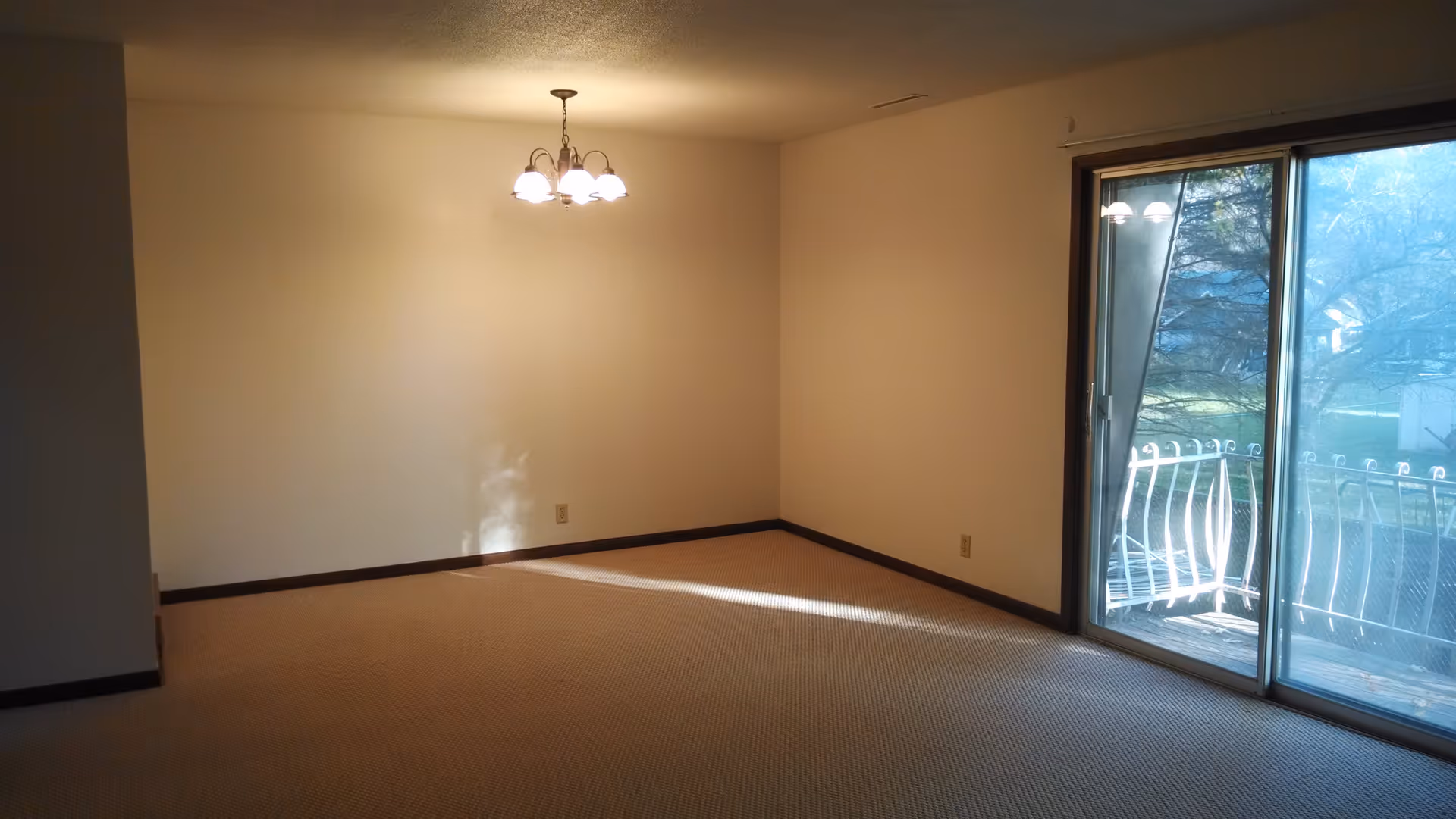 Empty carpeted living/dining room with a ceiling light fixture and a sliding glass door opening to a balcony.