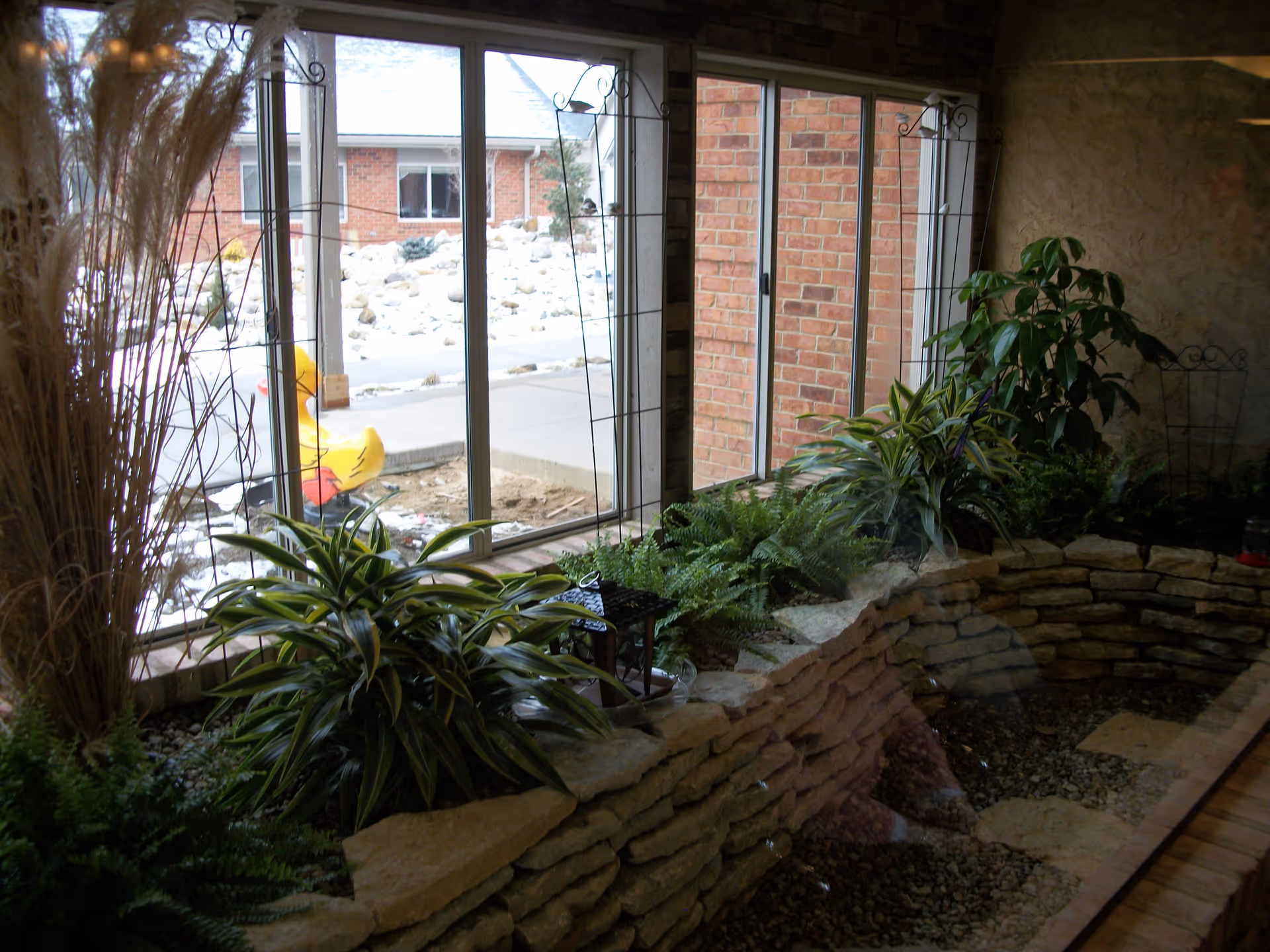 Indoor stone planter filled with green plants beside large windows overlooking a snowy courtyard and a yellow duck decoration.