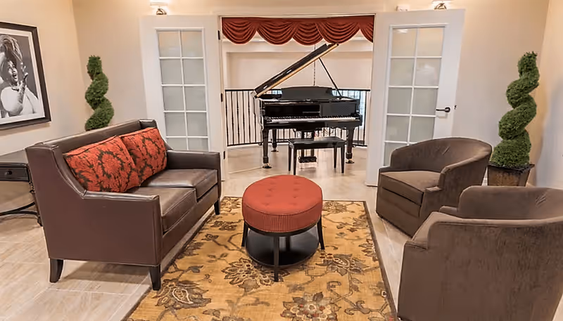 A cozy living room area with a brown leather sofa adorned with red patterned cushions, two brown upholstered armchairs, and a round red ottoman on a floral patterned rug. In the background, there is a black grand piano positioned in an adjacent room visible through open French doors. The room is decorated with two spiral-shaped topiary plants and a black and white framed photograph on the wall.