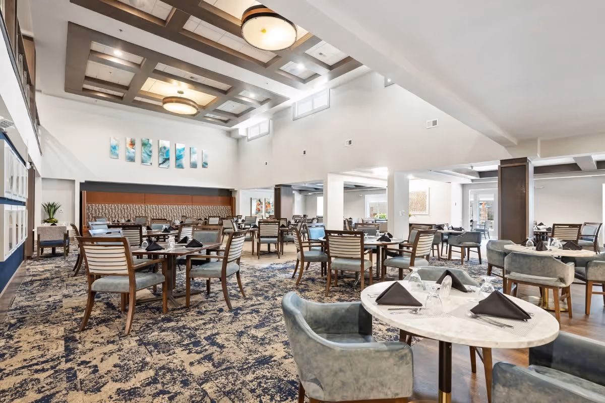A spacious and well-lit dining room with multiple tables and chairs arranged neatly. The tables are set with black napkins, glasses, and cutlery. The room features a patterned carpet, modern ceiling lights, and decorative artwork on the walls. Large windows near the ceiling allow natural light to brighten the space.