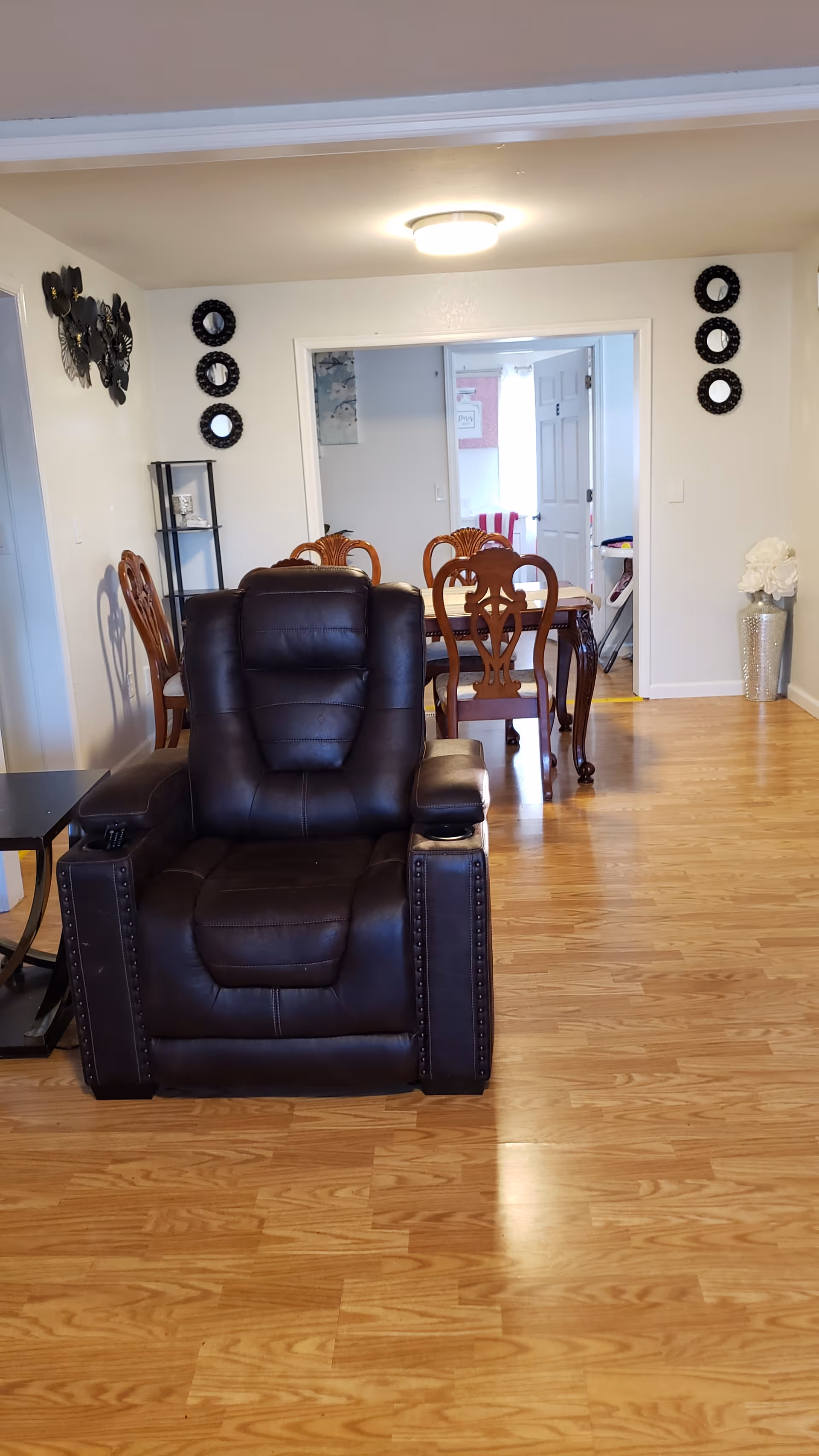 Interior view of a senior living facility showing a dark brown leather recliner chair in the foreground on a wooden floor. Behind the chair is a dining area with a wooden dining table and chairs. The walls are decorated with small round mirrors and a metal wall art piece. A ceiling light fixture illuminates the room.