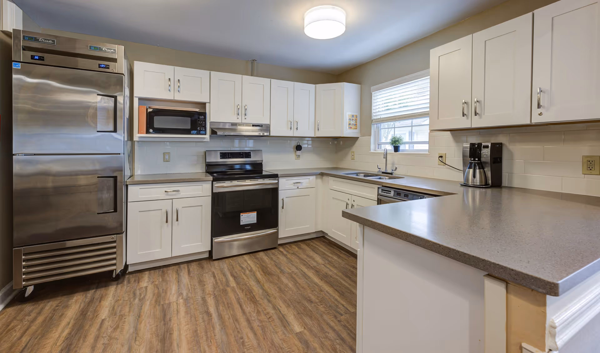 A clean, modern kitchen with white cabinets, stainless steel appliances including a large refrigerator, oven, microwave, and dishwasher. There is a coffee maker on the counter, a window with blinds above the sink, and wood-patterned flooring.