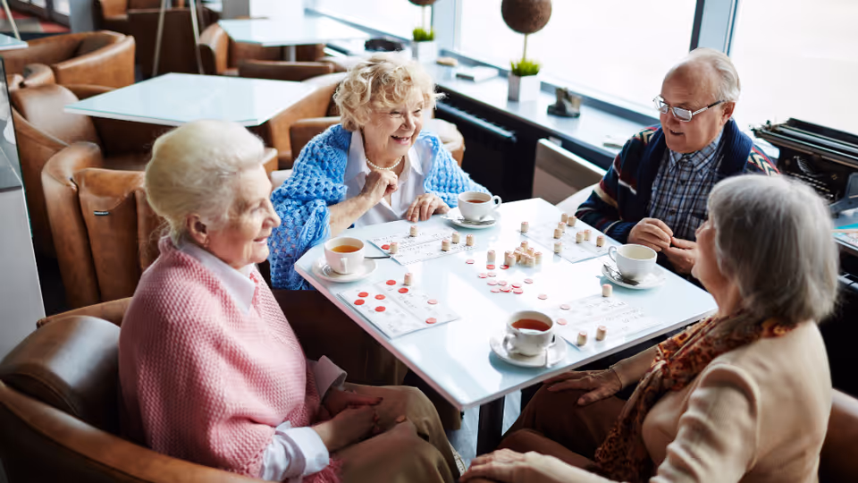Four elderly people sitting around a white table in a cozy lounge area, playing a game with bingo cards and markers, each with a cup of tea or coffee in front of them. The room has large windows letting in natural light and comfortable brown leather chairs.