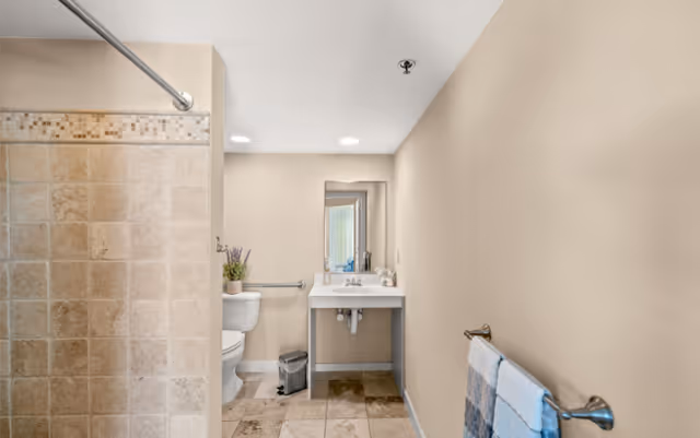 A clean bathroom with beige tiled floor and walls. On the left side, there is a tiled shower area with a metal shower curtain rod. Next to the shower is a white toilet with a small plant on the tank. In the center, there is a white sink with a mirror above it and a small trash can underneath. On the right wall, there is a towel rack holding two folded towels.