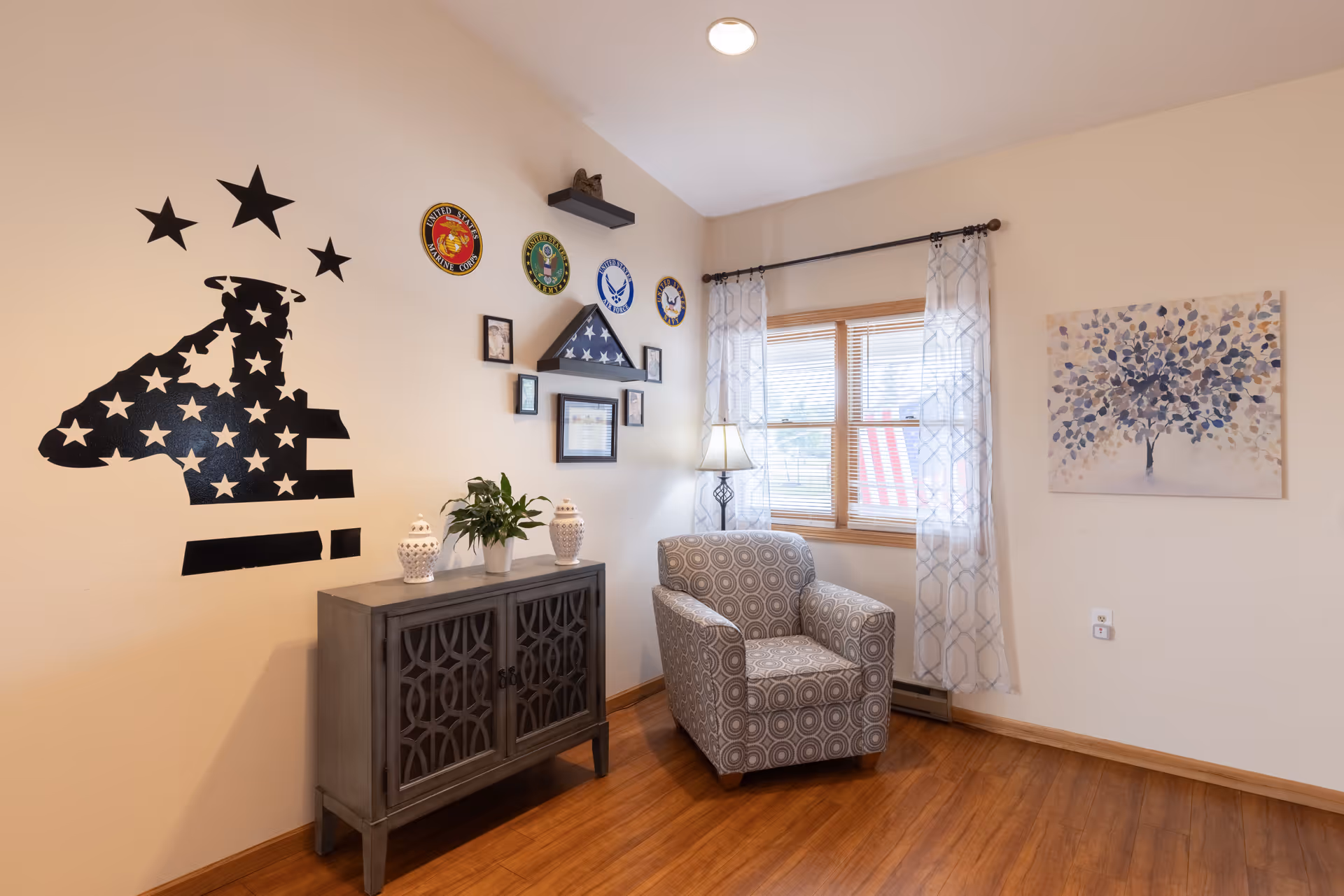 A cozy corner of a living room with a patterned armchair next to a window with sheer curtains. A small gray cabinet with decorative items sits against the wall, which is decorated with military insignia, framed photos, and a folded American flag in a display case. A large wall decal of a saluting soldier silhouette with stars is also visible.