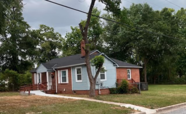 Single-story brick and light-blue siding house with a front porch and ramp surrounded by grass and trees.