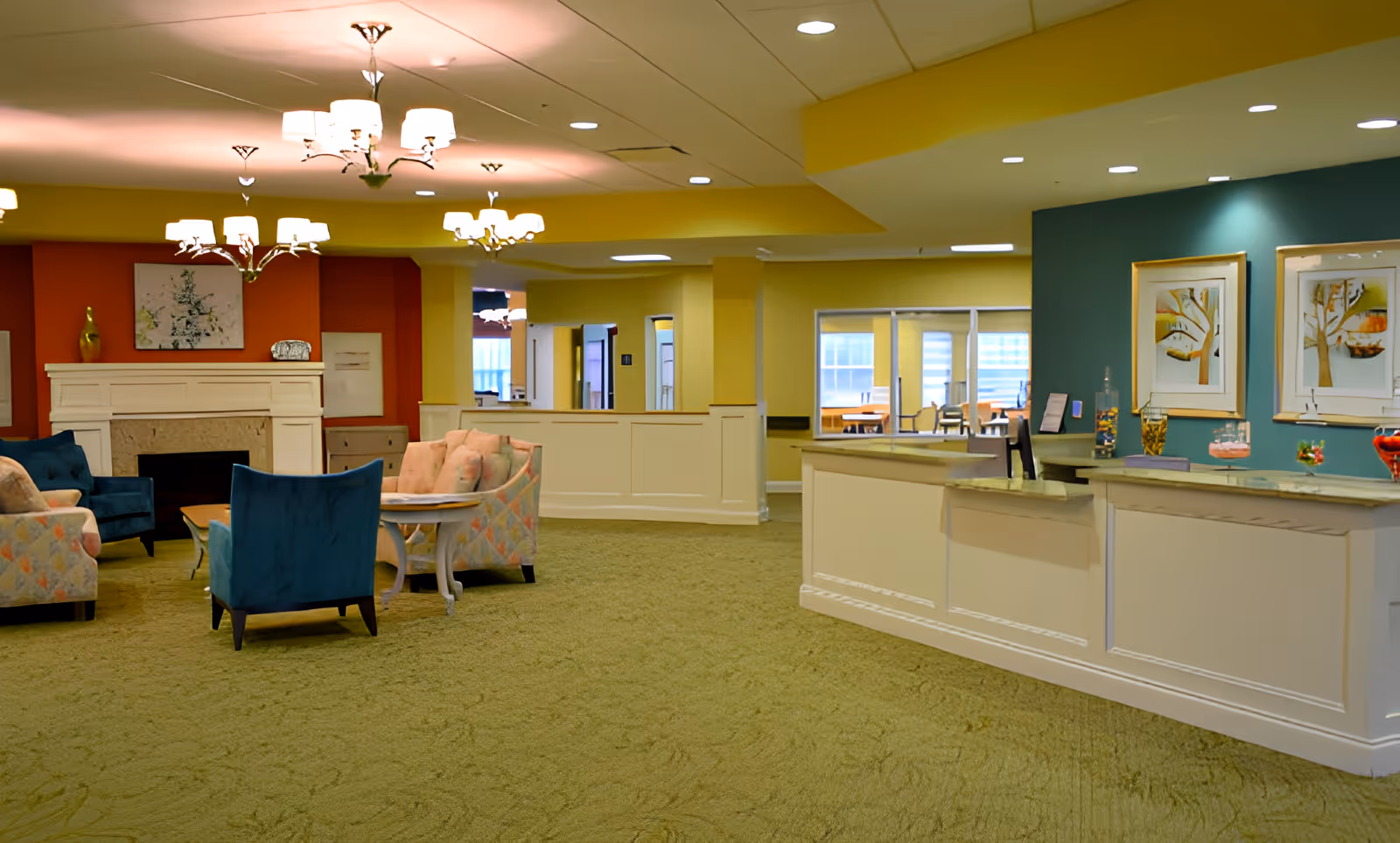 A spacious and well-lit common area in a senior living facility featuring a reception desk on the right with decorative items and framed artwork on the wall behind it. To the left, there is a seating area with upholstered chairs and sofas arranged around a coffee table in front of a fireplace with a red accent wall. The room has yellow walls, green carpet, and multiple ceiling light fixtures.
