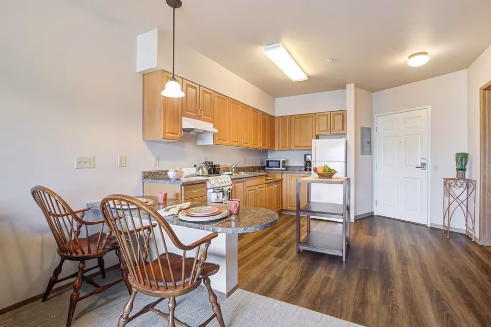 A bright kitchen and dining area with wooden cabinets, a white refrigerator, stove, microwave, and coffee maker. There is a small kitchen island with a bowl of fruit on it. Two wooden chairs are placed at a rounded countertop set for two with plates and mugs. The floor is wood laminate, and there is a white door and a small decorative table with a plant near the entrance.