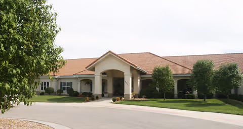 Exterior view of a single-story building with a tiled roof and an arched entrance, surrounded by green grass, trees, and shrubs under a partly cloudy sky.