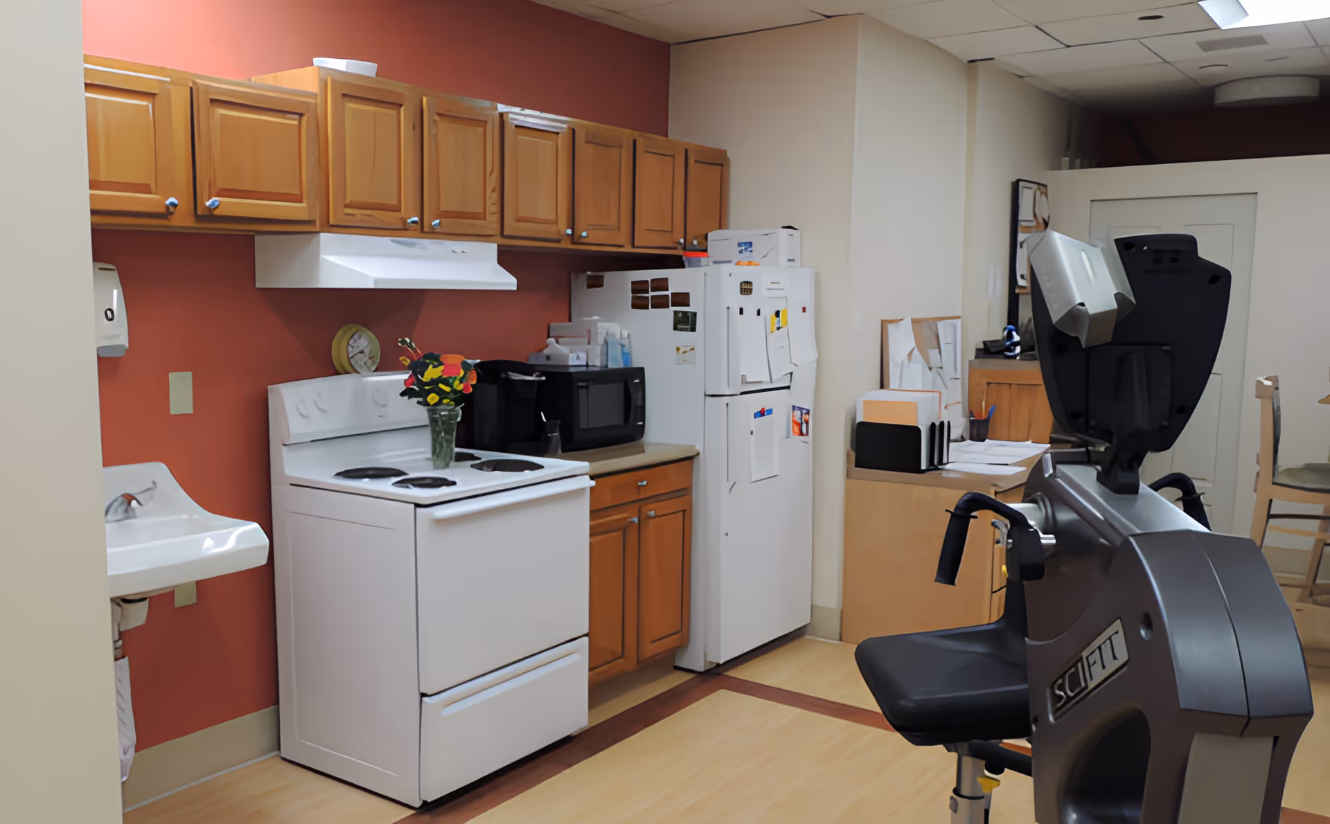Interior view of a room with a kitchen area featuring wooden cabinets, a white stove with a vase of flowers on top, a microwave, and a refrigerator covered with papers and magnets. To the right, there is a SCIFIT exercise machine and a small desk with office supplies and papers. The walls are painted in a combination of red and beige.