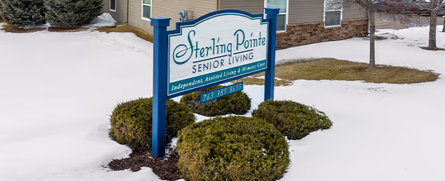 A sign for Sterling Pointe Senior Living stands outdoors on a snowy lawn with some green bushes around it. The sign reads 'Sterling Pointe Senior Living, Independent, Assisted Living & Memory Care' along with a phone number. A building with beige siding and stone accents is visible in the background.