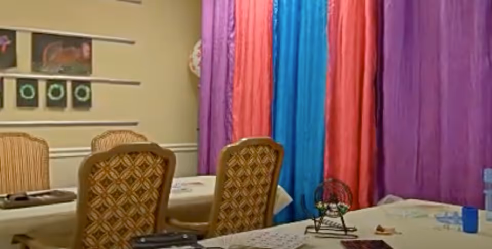 A room with tables and chairs arranged for a bingo game, featuring colorful curtains in purple, red, and blue hanging on the wall. There are bingo cards and a bingo cage on the tables, and framed artwork on the beige wall in the background.