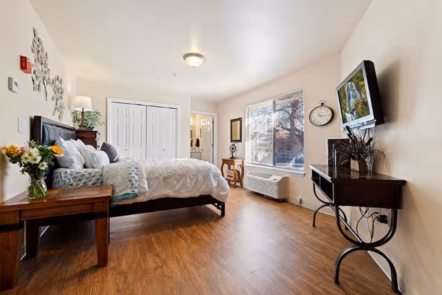 A bright and spacious bedroom with wooden flooring, a large bed with white and blue bedding, a wooden nightstand with a vase of flowers, a wall-mounted TV, a clock, and a window letting in natural light.